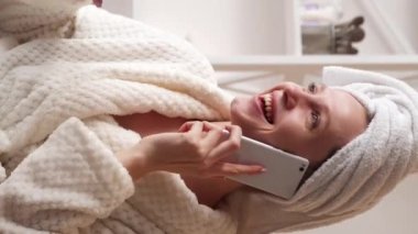 Dikey video. Kadın istirahatı. Dostça bir konuşma. Beyaz banyo spa havlusu ve duştan sonra telefonda sohbet eden rahat gülen genç kız..