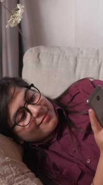 Dikey video. Akıllı telefon sohbeti. İnternet iletişimi. Ev tatili. Neşeli, gülümseyen bir kadın cep telefonundan mesaj atıyor rahat koltukta dinleniyor..