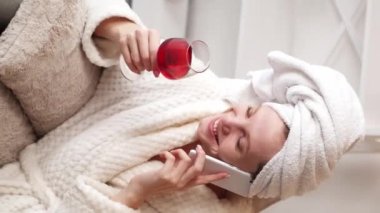 Dikey video. Şarap gevşetir. Sıcak bir sohbet. Beyaz banyo spa havlusu ve duştan sonra telefonda konuşan kırmızı camlı yumuşak bornozlu rahat gülen genç kız..