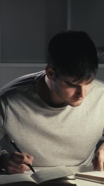 Dikey video. Test hazırlığı. Gece son teslim tarihi. Evde eğitim. Çalışkan erkek öğrenci kitap yazıyor karanlık kampüste geç saatlere kadar öğreniyor..