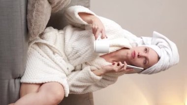 Dikey video. Ev spası. Bayan aradı. Beyaz banyo spa havlusu ve yumuşak bornoz giymiş genç bir kadın telefonda konuşuyor duştan sonra sıcak çay içiyor..