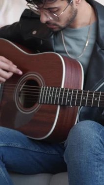 Dikey video. Gitar çalıyor. Yaratıcı hobi. İlham verici müzisyen akustik enstrüman tellerini akort ediyor evdeki kanepede ses kontrolü yapıyor..