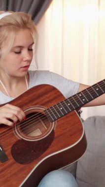 Dikey video. Gitar çalıyor. Profesyonel kadın. Ev dersi. Kulaklıklı ciddi bir kadın cep telefonu lambasını kullanarak enstrüman tellerini ayarlıyor..