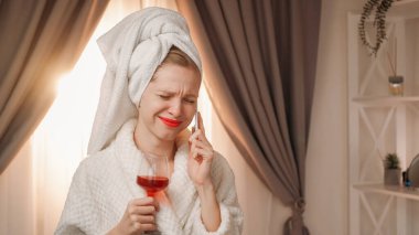 Wine tasting. Online discussion. Attractive young woman with makeup in white bath towel and bathrobe holding glass with red drink after shower while talking phone. clipart