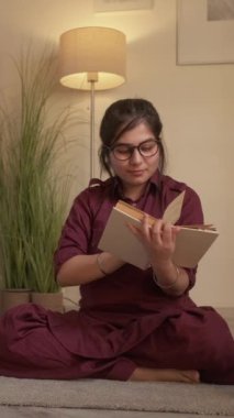 Dikey video. İyi akşamlar. Kitap hobisi. Bilgi yaşam tarzı. Sakin kadın öğrenci evde hikayeden zevk alıyor. Oturma odasında yerde ödev edebiyatı öğreniyor..