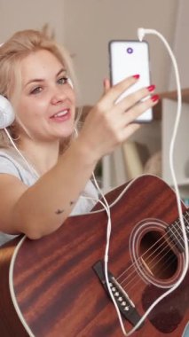 Dikey video. İnternetteki konferans. Mutlu kadın. Gitar çalıyor. Heyecanlı kulaklıklı bayan aydınlatma odasının içinde akıllı telefondan müzik aletini gösteriyor..