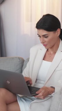 Dikey video. Ofis işleri. Zarif kadın. Dijital proje. İş kadını dizlerinin üzerinde dizlerinin üzerinde dizüstü bilgisayar yazıyor. İç mekânda koltukta oturuyor..