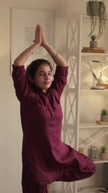 Dikey video. Yoga hobisi. Sağlıklı bir yaşam. Kapalı mekan eğlencesi. Vrikshasana ağacı pozu veren mutlu ve rahat genç bir kadın evde dikkatli hareketlerin tadını çıkarıyor..