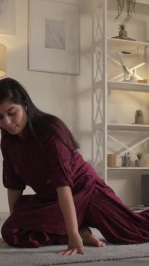 Dikey video. Yoga egzersizi. Evde eğitim. Kapalı alan aktivitesi. Esnek, neşeli, sağlıklı bir kadın esneyerek tek bacaklı güvercin pozu egzersizi yapıyor oturma odasında..