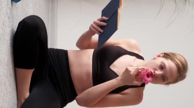Dikey video. Bayan sakin olun. Spor arası. Harika fitness kızı siyah takım elbise giyip kitap okurken lezzetli pastalar yiyor..