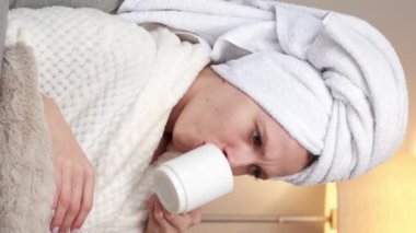 Dikey video. Sıvı içecek. Spa uykusu. Beyaz banyo havlusu ve yumuşak bornozlu rahat genç kız duştan sonra sıcak kahve içiyor..