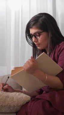 Dikey video. Edebiyat okuyorum. Evde eğitim. Sınav hazırlığı. Konsantre olmuş genç bir kadın günlüğe ödev notları yazıyor rahat koltukta kitap öğreniyor..