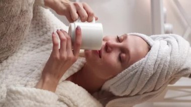Dikey video. Spa tedavisi. Çay gevşemesi. Banyo havlusu ve beyaz yumuşak bornoz giymiş, duştan sonra sıcak içecek içen sakin bir kadın..