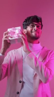 Dikey video. Romantik hediye. Sevgililer günü hediyesi. Tatil sürprizi. Neon rengi açık renk, neşeli, gülümseyen, gözlüklü, pembe kutuda izole edilmiş yakışıklı adam..