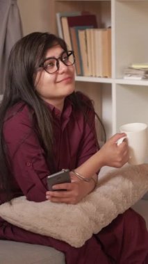 Dikey video. Kahve molası. Kapalı alanda dinleniyor. Gezici eğlence. Mutlu rüya gibi kadın sıcak bir bardak içki içiyor akıllı telefon dinleniyor evdeki oturma odasında kanepede dinleniyor..