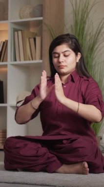 Dikey video. İçerde meditasyon. Yoga sağlığı. Farkındalık pratiği. Gyan mahamayuri dhyana mudra jesti yapan barışçıl kadın evinde, oturma odasında nilüfer pozisyonunda.