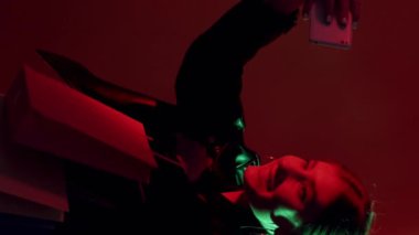 Dikey video. Alışveriş selfie 'si. Kara Cuma. Gece satışı. Kırmızı yeşil renkli, neon ışıklı, çantaları olan, kameraya poz veren, koyu renk öpücüklü kız..