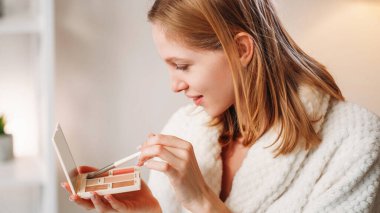 Makyaj zevki. Cilt bakımı. Rahat kız beyaz bornozlu toz fırçası kullanarak makyaj yapıyor. Aynada yansımasının tadını çıkarıyor..