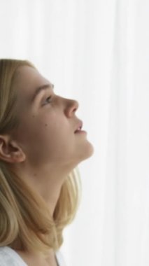 Dikey video. Sabah başlıyor. Motivasyon anı. Gün ışığı enerjisi. Olumlu, sakin bir kızın perdeleri açması, gülümsemesi, ferahlatıcı düşüncelere dalması, pencereden bakması..