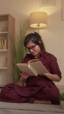 Dikey video. Kitap eğleniyor. Bilgi ve rahatlık. Evde dinleniyorum. Oturma odasında, yerde oturmuş kitap okuyan mutlu, zeki bir kadın öğrenci..