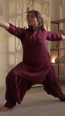 Dikey video. Yoga esneme hareketleri. Sağlıklı aktivite. Kapalı alanda eğitim. Virabhadrasana savaş güvercini pozu veren motive olmuş bir kadın. Evdeki refahın tadını çıkarıyor..