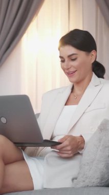 Dikey video. İş toplantısı. Zarif kadın. İnternetteki konferans. Laptopta konuşan kendine güvenen bir kadın. İç mekânda koltukta oturuyor..