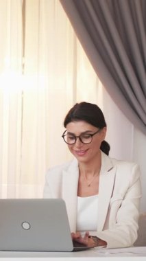 Dikey video. Ofis işleri. Pozitif kadın. Sanal sohbet. Şık takım elbiseli mutlu bayan, iç mekânda dizüstü bilgisayar oturuyor..