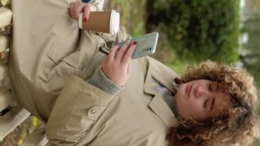 Dikey video. Gadget 'ın boş vakti. Mobil sohbet. Rahatlamış, huzurlu, tombul bir kız. Kahveli. Sonbahar parkında bankta mesajlaşarak eğleniyor..