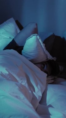 Dikey video. Gece uykusuzluğu. Uyku sorunu. Huzursuz adam yatakta dönüp başını yastıkla örtüyor loş mavi ışığın altında.