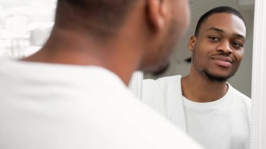 Erkek bakımı. Deri rutini. Sabah hijyeni. Taze yüzlü, kendine güvenen adam. Cilt beyazı. Işıksız arka planda aynaya bakıyor..