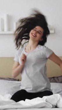 Dikey video. İyi sabahlar. Hafta sonu tatili. Dinlenmiş enerjik kadın rahat yatak odasında dans ediyor..