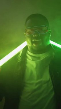 Dikey video. Rap şovu. Gece kulübü performansı. Havalı, kendine güvenen, modaya uygun güneş gözlüklü neşeli bir adam hip hop etkinlik sahnesinde parlak neon lambalı arka planda şarkı söylüyor..