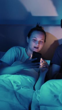 Dikey video. Genç çift gece lambasıyla aydınlatılan akıllı telefon dizüstü bilgisayarında sohbet ediyor..