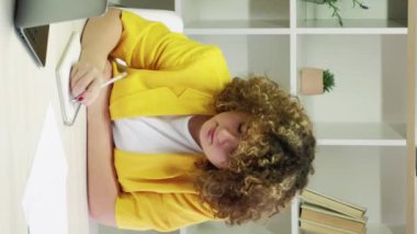 Dikey video. Mesafe öğrenme. Çevrimiçi çalışma. Uzaktan eğitim. Kıvırcık saçlı tombul öğrenci ev içi aydınlık ofis içi bilgisayar dersi için not alıyor..