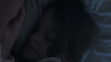 Dikey video. Uyuyan çocuk. Gece uykusu. Tatlı rüyalar. Çocuk yatma vakti. Şirin, küçük bir kız, rahat, yumuşak bir yatakta uzanıyor. Evdeki karanlık odayı mavi ay ışığıyla çeviriyor..