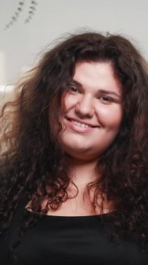 Dikey video. Doğal güzellik. Gülen surat. Vücut pozitif. Kendini kabullenme. Neşeli, güler yüzlü, kendine güveni tam, şişman, temiz tenli, siyah kıvırcık saçlı bir kadın..