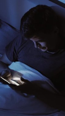 Dikey video. Karanlık yatak odalı adam gece yatağında uzanıyor. Akıllı telefon kullanıyor. Mavi ışıkla aydınlatılıyor..