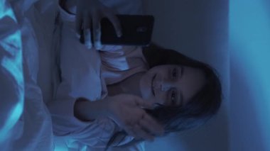 Dikey video. Gece çevrimiçi arama. Çocuk sohbeti. Geç iletişim. Neşeli küçük kız cep telefonu kullanıyor. El sallıyor. Evdeki karanlık odada mavi ışıkla oturuyor..