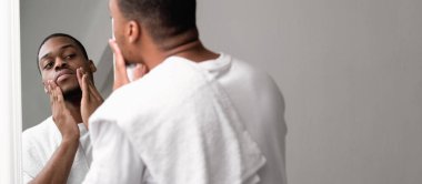 Deri rutini. Sakal bakımı. Beyaz havluyla kendine güveni tam bir erkek. Aynaya bakıyor. Boş alan arka planında yüz kıllarına dokunuyor..