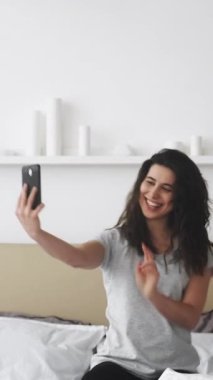 Dikey video. Selfie eğlencesi. Milenyum kadını. Çevrimiçi blog yazarı. Akıllı telefondan fotoğraf çeken mutlu bir kadın poz veriyor. Yumuşak yatak aydınlığında rahat bir yatak odasında oturuyor..