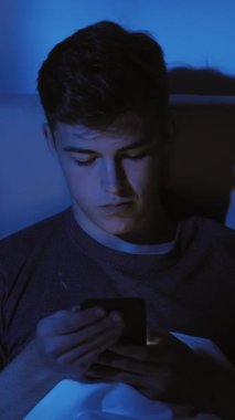 Dikey video. Genç adam, loş aydınlatılmış odada akıllı telefon kullanmaya daldı. Teknoloji yalnızlığı kavramlarını vurguladı..