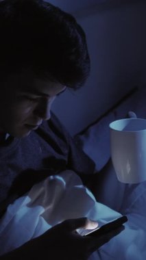 Dikey video. Karanlık odada uyuyan adam yatakta akıllı telefon fincanı kullanmadan önce rahatlama bağlantısı kurar ve rahat bir gece rutini yaratır..
