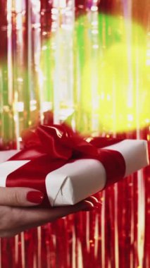 Dikey video. Tatil sürprizi. Noel hediyesi. Kadın eller, parlak kırmızı arkaplanda renkli bokeh ışığı tutuyorlar..
