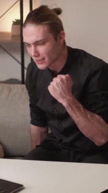 Dikey video. İş başarısı. İş haberleri. Siyah gömlekli genç adam masada oturup dizüstü bilgisayardaki e-postaları okuyunca mutlu oluyor..