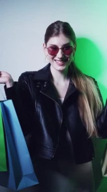 Dikey video. Alışveriş satışı. Kara Cuma. Alışverişkolik yaşam tarzı. Yeşil neon ışıklı mutlu, neşeli, gözlüklü ve çantalı çekici kız satın almanın keyfini çıkarıyor..