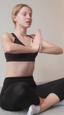 Dikey video. Yoga keyfi. Meditasyon eğlencesi. Siyah spor kıyafetli genç, zayıf, güzel bir kadın beyaz arka planda gözlerini kapatarak spor yapıyor..