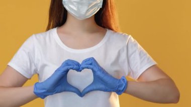 Aşk sinyali. Karantina ilişkisi. Korunaklı yüz maskeli kadın. Eldivenler, sarı fotokopi alanında izole edilmiş kalp hareketi gösteriyor..