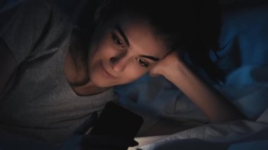Gezici gece. Gadget uykusuzluğu. Rahatlamış gülümseyen kadın telefon uygulamasını kullanmaktan zevk alıyor. Karanlık ev yatak odasında yatakta kaydırma yapıyor..