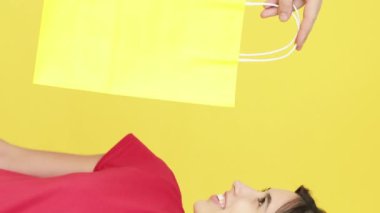 Dikey video. Hediye sürprizi. Beklenmedik bir hediye. Heyecanlı neşeli kadın sarı boş alanda izole edilmiş alışveriş torbaları alıyor..