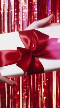 Dikey video. Tatil hediyesi. Mutlu Noeller. Kadın elleri, parlak arkaplanda kırmızı kurdeleli hediye kutusunu gösteriyorlar..